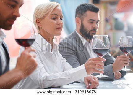 Serious blonde woman evaluating color of red wine in bokal by workplace