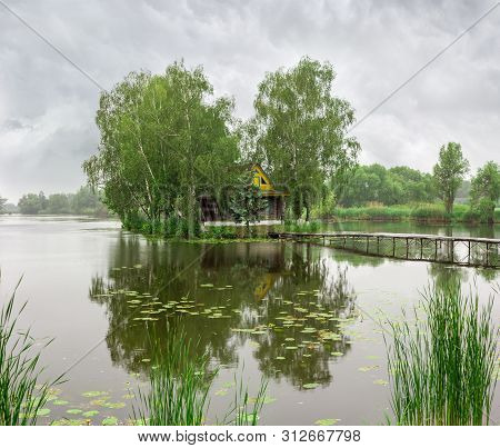 Tiny Island With Trees And Romantic Fishing Hut On Lake And Small Wooden Footbridge To Him In Summer