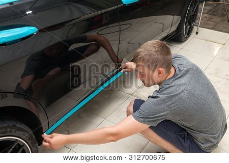 A Man Of Caucasian Appearance With Blue Paper Tape Plasters A Body Element Doorsill With A Black Car