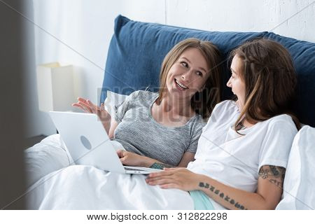 Two Smiling Lesbians Looking At Each Other While Using Laptop In Bed In Morning