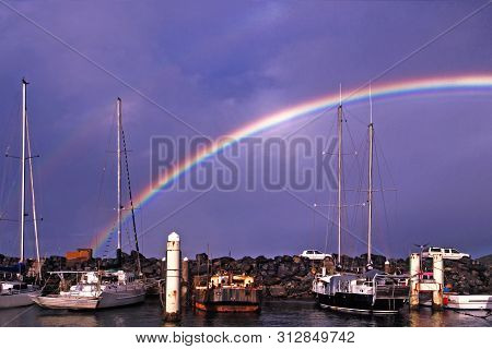 Rainbow In A Dark Blue Sky Over Waterfront Maritime Marina/dock With Berthed Boats Against A Rock Br