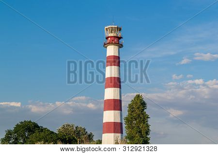 Lighthouse On The Beach On A Sunny Day Against The Blue Sky.  Trees Grow Near The Lighthouse.