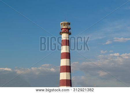 Lighthouse On The Beach On A Sunny Day Against The Blue Sky.  Trees Grow Near The Lighthouse.
