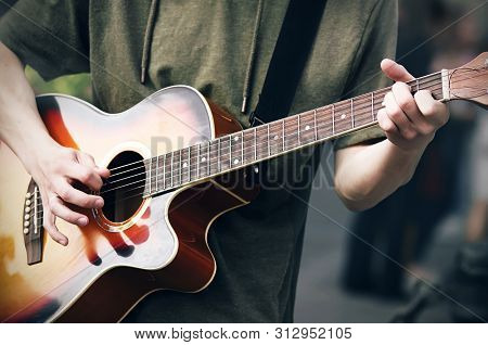 A Young Cocky Guy In A Green Hoodie Plays A Bright Colored Acoustic Six-string Guitar, Holding The C