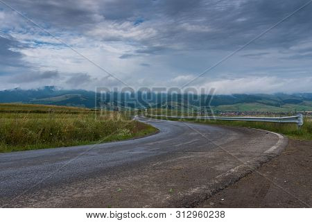 Empty, Curved Asphalt Road After Rain , Stormclouds On The Sky, Village In The Background In Transyl