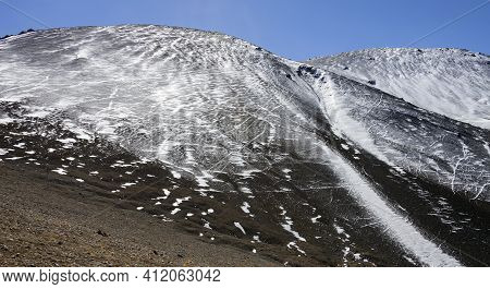 Background Dark Mountain Sprinkled With White Snow With Trails And Footprints. A Rocky, Loose Mounta