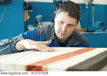 A Cabinetmaker Works With Wood In A Carpentry Shop. A Young Worker Of Caucasian Appearance In A Unif