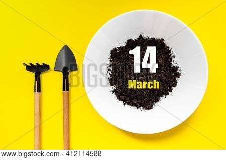 March 14th. Day 14 Of Month, Calendar Date. White Plate Of Soil With A Small Spatula And Rake On Yel