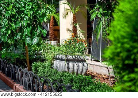 Stone Flower Pot With Flowers In A Flower Bed With Bushes And Trees Sprinkled With Tree Bark Mulchin