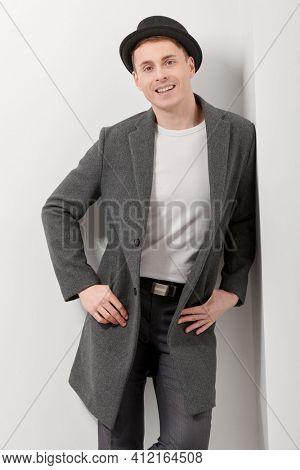 Porttrait of a handsome middle-aged man in an elegant coat and a hat standing by a white wall and smiling. Men's beauty, fashion. Lifestyle. Hipster man. 
