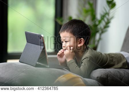 Asian Boy Looking At The Tablet Screen Attentively. Overstimulated Children Concept. Too Much Screen