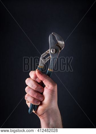 A Raised Hand Of Master Plumber Holds Pliers Against A Dark Background. Sign Of Successful Completio
