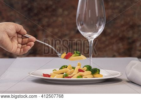 Selective Focus Of A Fork With Pasta And Vegetables Held Bu A Hand Over A Plate Of Pasta Salad On An
