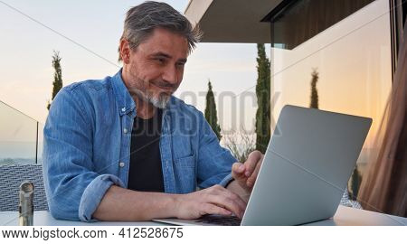 Happy man working online with laptop computer on terrace. Businessman in home office, browsing internet, tele working, video chat.