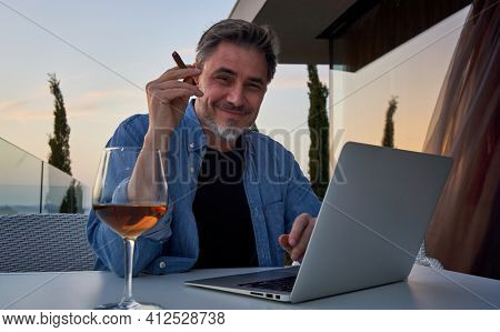 Man relaxing at home. Man sitting on balcony using laptop, enjoying wine and cigar. Home office, tele working, lifestyle, escape, freedom, getaway.