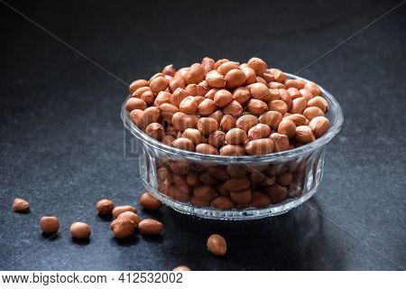 Stock Photo Of A Brown Color Raw Peanuts In Transparent Glass Bowl, Kept On Black Granite Stone Unde