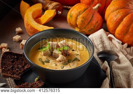 Pumpkin cream soup with pumpkin seeds and dried bread cubes. Lenten menu. Healthy, vegetarian food. Bowl with soup  and orange pumpkins on gray background.