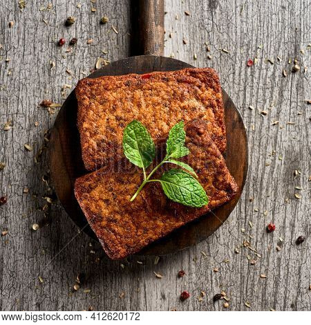 high angle view of some cooked tofu fillets in a round wooden tray, placed on a gray rustic wooden table sprinkled with different herbs and spices
