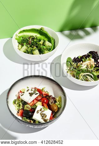Greek salad, cheese and beet salad and cucumbers with chuka seaweed. Salad bowl on white table with green wall. Sunlight with harsh shadow of green tropic leaf