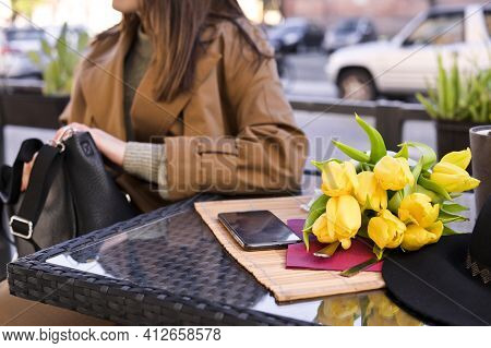 Young Beautiful Girl In A Cafe On The Street. Spring Sun In The Frame. A Bouquet Of Flowers Lies On 