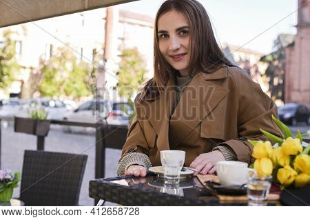 Young Beautiful Girl Drinks Espresso In A Cafe On The Street. Spring Sun In The Frame. A Bouquet Of 