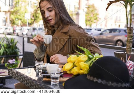 Young Beautiful Girl Drinks Espresso In A Cafe On The Street. Spring Sun In The Frame. A Bouquet Of 