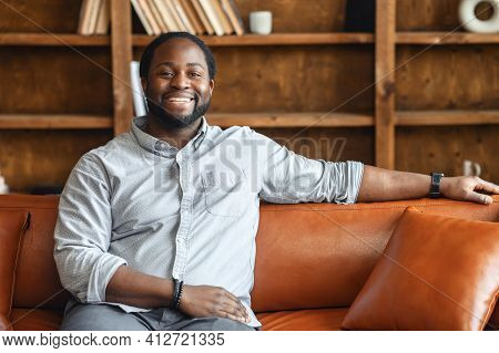 Ethnic Black Unshaved Plump Man With Dreadlocks Sitting On The Leather Sofa And Smiling, In A Normal