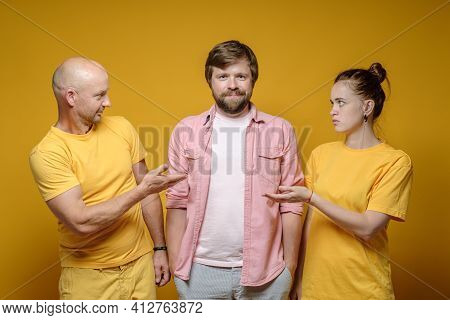 Man And A Woman Look At Their Contented And Smiling Friend In Bewilderment. Yellow Background.