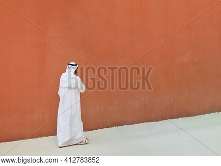 Arabic Man With Traditional Emirates Clothes Walking Outdoors In The Street Against A Orange Wall Ba