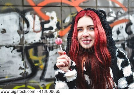 Portrait Of A Young Beauty In A Hoodie With A Delicious Pink Lollipop, Posing Against A Wall Of Graf