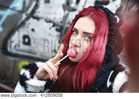Portrait Of A Young Beauty In A Hoodie With A Delicious Pink Lollipop, Posing Against A Wall Of Graf