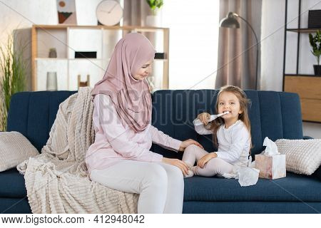 Young Mother, Pretty Muslim Woman In Hijab And Light Domestic Clothes, Sitting On Blue Sofa At Home 
