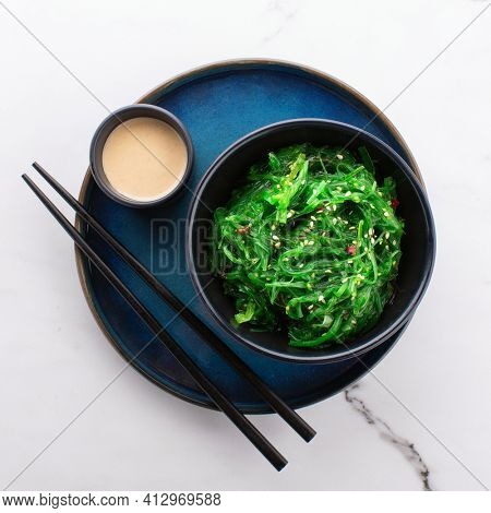 Top view of healthy green seaweed salad in a black bowl, peanut sauce and chopsticks on a beautiful blue plate on a white stone background.