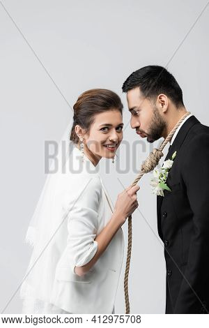 Cheerful Bride Holding Slip Knot On Neck Of Arabian Groom Pouting Lips Isolated On Grey.