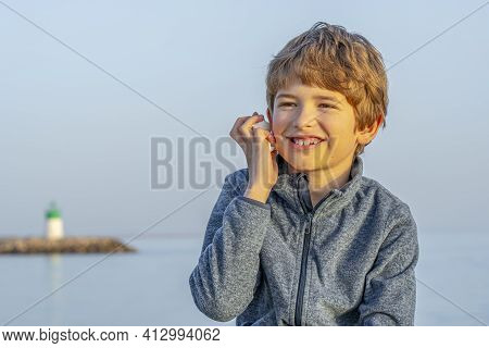 Close Up Portrait Of A Cute Young Boy Holding A Sea Shell To His Ear. Happy Boy Listening To The Sou
