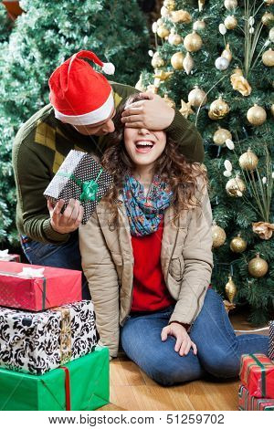 Young man in Santa hat surprising woman with Christmas gifts in store