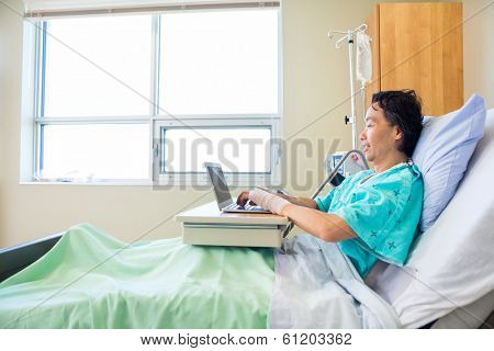 Full length side view of mature male patient using laptop on bed in hospital