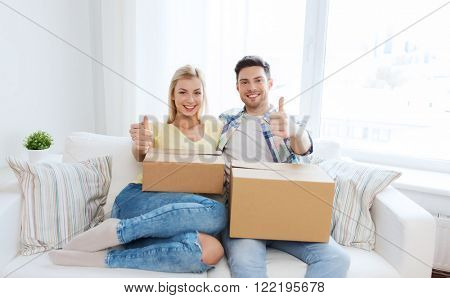 moving, people, repair and real estate concept - happy couple with big cardboard boxes showing thumbs up on sofa at new home