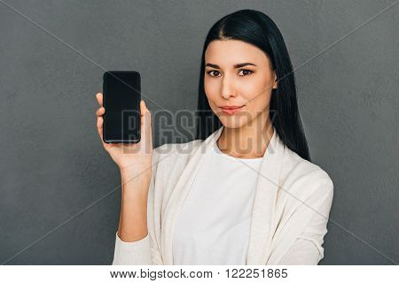 Take a look here! Beautiful young woman shoving smartphone with copy space and looking at camera while standing against grey background