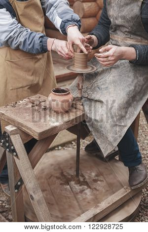 The hands of a potter help make a pitcher on a pottery wheel. Two men with hands caked in clay. One man teaches the second pottery.