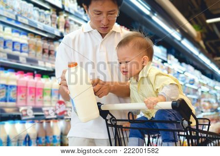 Asian Father & Cute little 18 months / 1 year old toddler boy child choosing milk product in grocery store, Dad read milk nutrition facts with child sit in shopping cart, Kid first experience concept