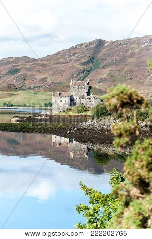 Majestic castle at Eilean Donan reflected in the high tide on Loch Alsh with sunny mountains and blue sky on background. Summertime outdoors vertical image.