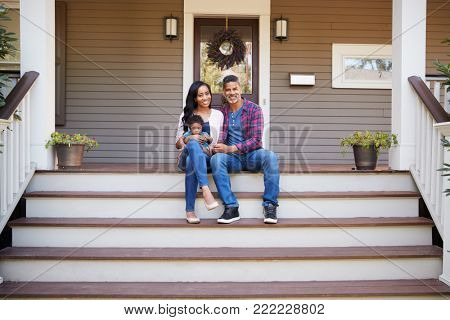 Family With Baby Son Sit On Steps Leading Up To Porch Of Home