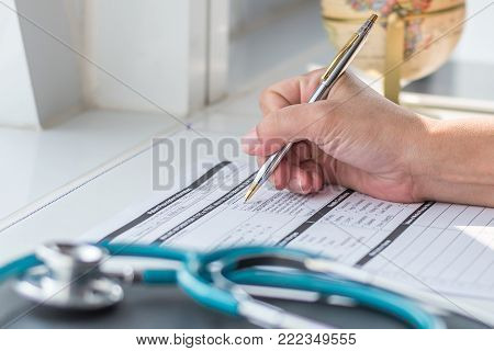 Doctor writing on medical health care record, patients discharge, or prescription form paperwork in hospital clinic office with physician's stethoscope on desk