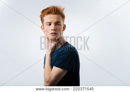 Preoccupied. Handsome humourless red-headed young man touching his neck and wearing a black T-shirt and thinking