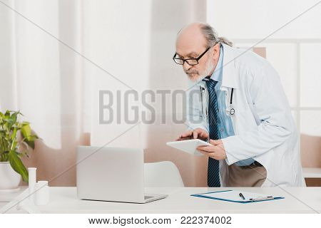 portrait of senior doctor in white coat with tablet in hands at workplace in clinic