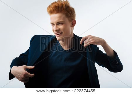 Happiness. Attractive content red-haired young man putting his jacket on and smiling and looking in the distance
