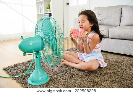 attractive young female little children sitting in front of electric fan blowing cooling wind and eating cold watermelon feeling refreshing during summer season.