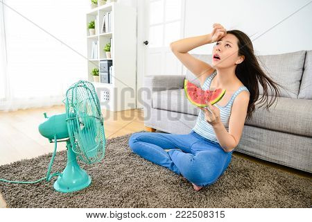 pretty elegant woman blowing electric fan with eating watermelon to refreshing when she feeling hot and getting headache painful at home in summer season.