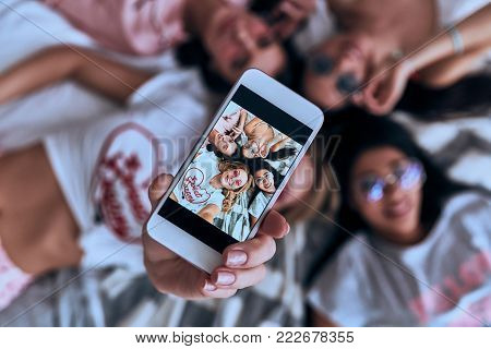 Perfect selfie. Top view of four playful young smiling women in eyewear taking selfie with smart phone while lying on the bed at home